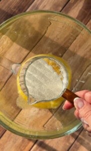 A hand holds a measuring cup of granulated sugar over a large glass bowl filled with melted butter, ready to pour it in. The scene is set on a wooden surface.