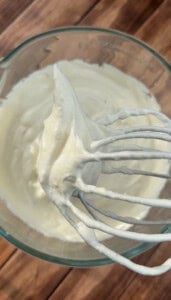 A metal whisk with stiff peaks of whipped cream is held above a glass bowl filled with more whipped cream, set on a wooden surface.