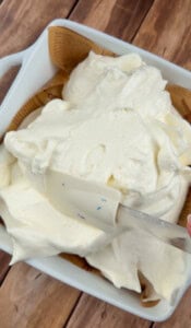 A close-up of a white baking dish filled with fluffy whipped cream being spread with a clear silicone spatula, set on a wooden surface.