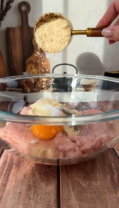 A hand pours breadcrumbs from a measuring cup into a glass bowl containing ground chicken, an egg, salt, and pepper, on a wooden surface with kitchen items in the background.