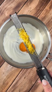 A hand holds a grater with fresh lemon zest over a metal bowl containing two eggs and white liquid, likely yogurt or cream, on a wooden surface.