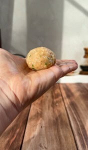A close-up of a hand holding a round, uncooked dough ball with herbs, above a wooden surface in natural sunlight.