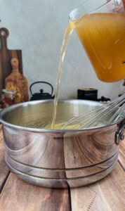 A hand pours golden broth from a glass measuring cup into a stainless steel pot containing a whisk, set on a wooden countertop with kitchen utensils in the background.