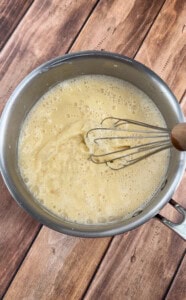 A metal whisk stirring a creamy, light yellow mixture in a stainless steel pot on a wooden surface.