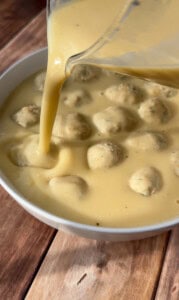 Creamy yellow sauce is being poured from a blender over a bowl filled with round dumplings or meatballs, all placed on a wooden surface.