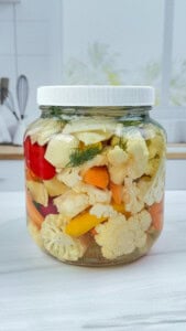 A glass jar filled with assorted pickled vegetables, including cauliflower, carrots, peppers, and herbs, sits on a white countertop in a bright kitchen setting.