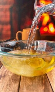 A stream of hot water is being poured from a glass pitcher into a clear bowl containing sliced lemons, with a warm, cozy fireplace and a mug in the blurred background.
