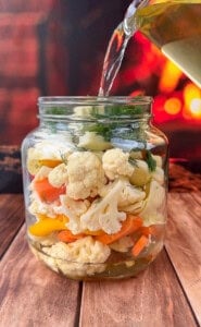 A glass jar filled with cauliflower, carrots, yellow peppers, and herbs is being covered with a clear liquid, likely for pickling, on a wooden surface with a warm, blurred background.