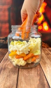 A hand placing sliced carrots into a glass jar filled with assorted chopped vegetables, including cauliflower and yellow peppers, on a wooden surface with a blurred warm, fiery background.