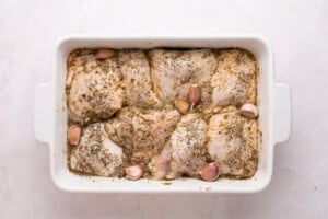 Raw chicken thighs seasoned with herbs and spices, arranged in a white rectangular baking dish with whole garlic cloves, ready for baking.