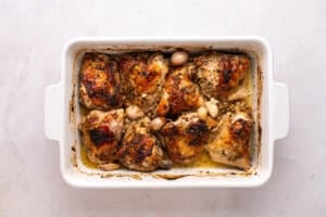 Baked chicken thighs with a crispy, golden-brown skin and whole garlic cloves in a white rectangular baking dish with juices visible at the bottom, on a light marble surface.