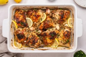 Golden-brown roasted chicken thighs in a white baking dish, garnished with lemon wedges, whole garlic cloves, and chopped herbs. The dish is on a white surface with a beige napkin and parsley nearby.