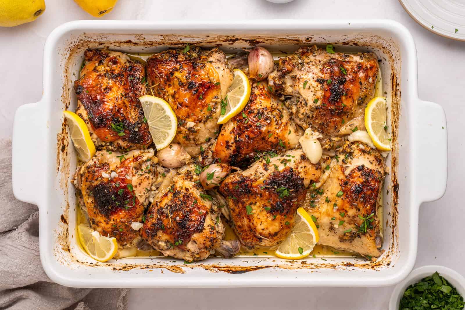Golden-brown roasted chicken thighs in a white baking dish, garnished with lemon wedges, whole garlic cloves, and chopped herbs. The dish is on a white surface with a beige napkin and parsley nearby. - 11