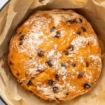 A round loaf of chocolate chip bread with a golden, lightly floured crust sits in a parchment-lined pot, ready for baking.