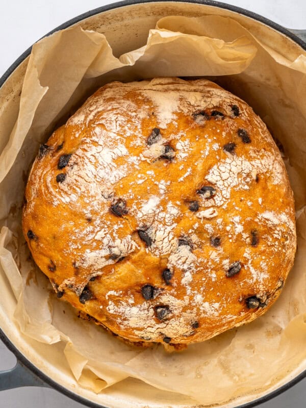 A round loaf of chocolate chip bread with a golden, lightly floured crust sits in a parchment-lined pot, ready for baking.