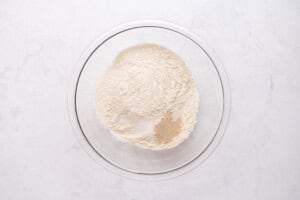 A clear glass bowl containing a mixture of dry ingredients—flour, yeast, salt, and sugar—on a white marble surface.