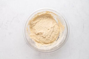 A glass bowl containing pale, thick batter sits on a white marble surface. The batter appears to be well-mixed, with some residue on the sides of the bowl.