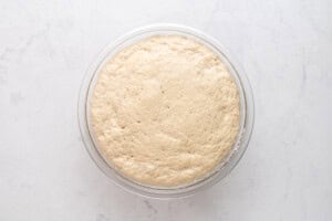 A glass bowl filled with risen, bubbly bread dough sits on a white marble surface, viewed from above.