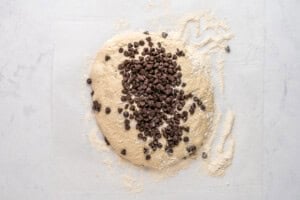 Chocolate chip cookie dough on a floured surface, topped with a generous amount of chocolate chips, ready to be mixed and shaped for baking.