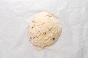 A round ball of bread dough with visible chocolate chips is placed on a sheet of parchment paper. The dough appears soft and slightly floured.