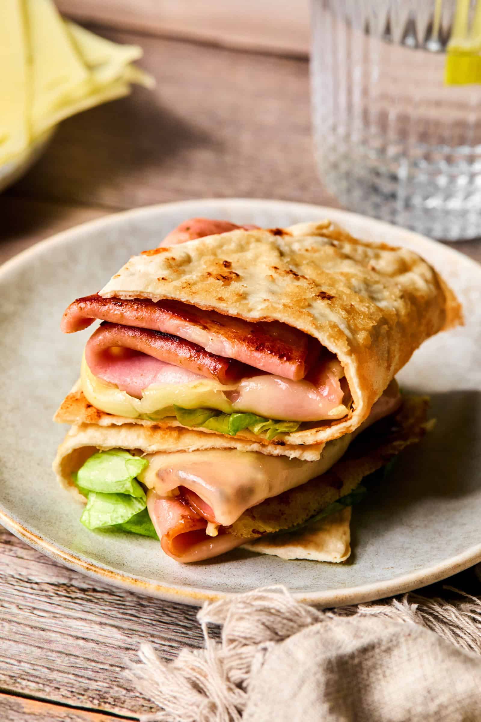 A close-up of Low Carb Mortadella Wraps, folded flatbread stuffed with melted cheese, ham, and lettuce, stacked on a beige plate. A glass of water and slices of cheese sit in the background on a wooden table.