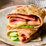 A close-up of two folded Low Carb Mortadella Wraps filled with lettuce, slices of ham, cheese, and sauce, stacked on a beige plate with a glass of water in the background.