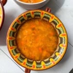 A decorative bowl filled with vegetable soup, featuring visible pieces of carrot, cabbage, and herbs, placed on a white wooden table next to another bowl of soup.