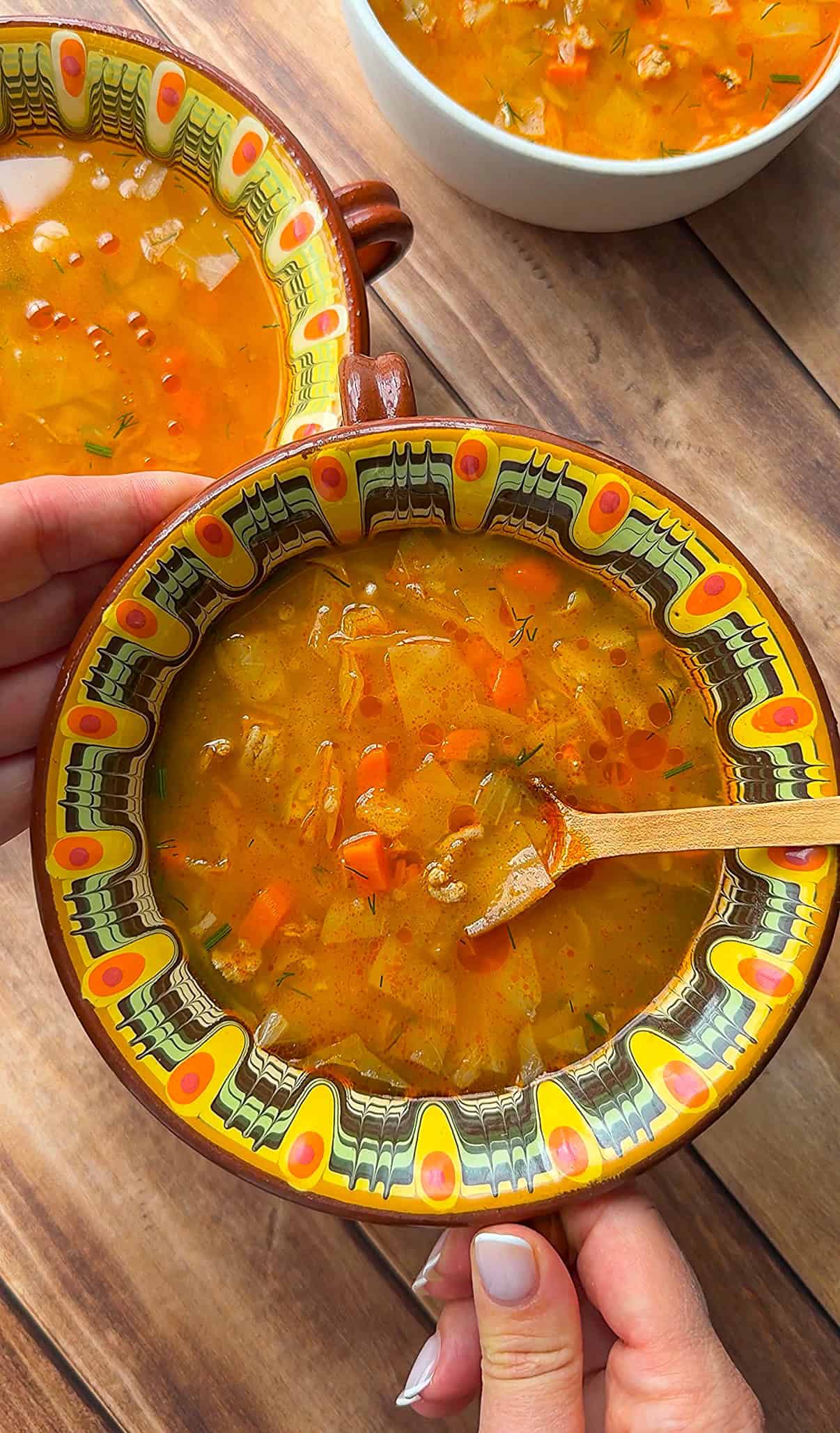 A hand holds a colorful, patterned bowl filled with vegetable soup, with another hand stirring using a wooden spoon. Two more bowls of similar soup are visible on a wooden table in the background.