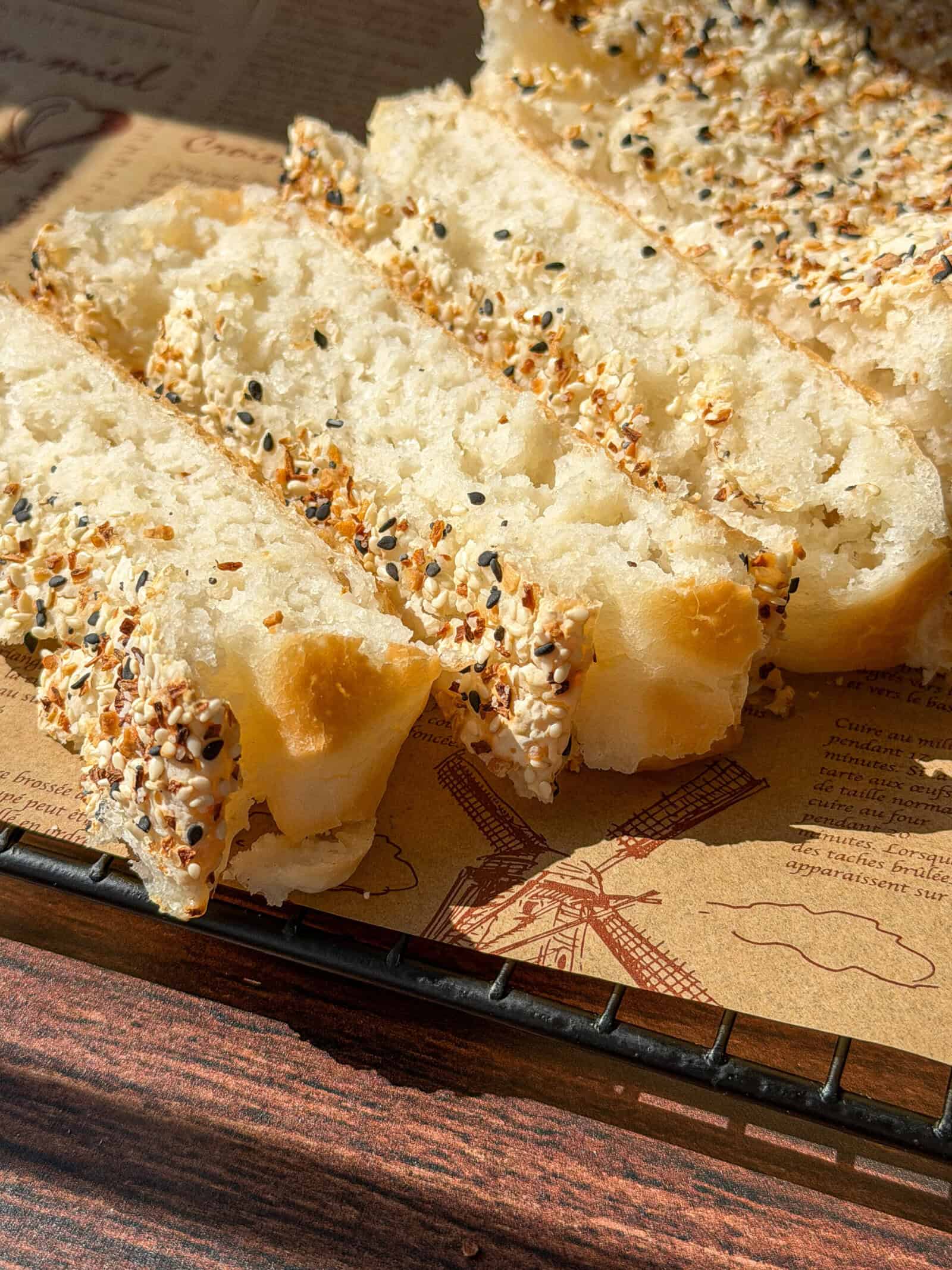 Sliced Viral Blender Rice Bread topped with a mix of sesame seeds and seasoning sits on parchment paper printed with drawings, including a windmill, on a wire cooling rack in sunlight.