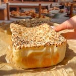 A hand holds a rectangular, seeded bread—reminiscent of the viral Blender Rice Bread—covered in sesame and poppy seeds, resting on parchment paper with a bakery display of breads and pastries in the background.