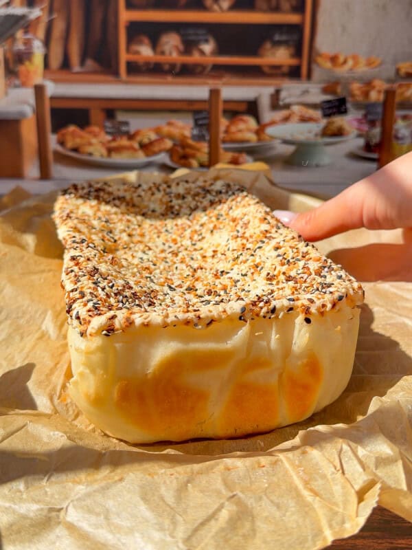 A hand holds a rectangular, seeded bread&mdash;reminiscent of the viral Blender Rice Bread&mdash;covered in sesame and poppy seeds, resting on parchment paper with a bakery display of breads and pastries in the background.