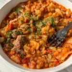 A close-up of a bowl filled with a hearty vegetable and meat stew, topped with fresh chopped herbs. A wooden fork rests inside the bowl, ready for eating. The dish appears thick, with visible chunks of meat, cabbage, carrots, and tomatoes.