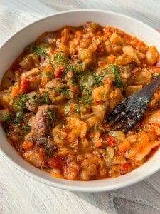 A close-up of a bowl filled with a hearty vegetable and meat stew, topped with fresh chopped herbs. A wooden fork rests inside the bowl, ready for eating. The dish appears thick, with visible chunks of meat, cabbage, carrots, and tomatoes.