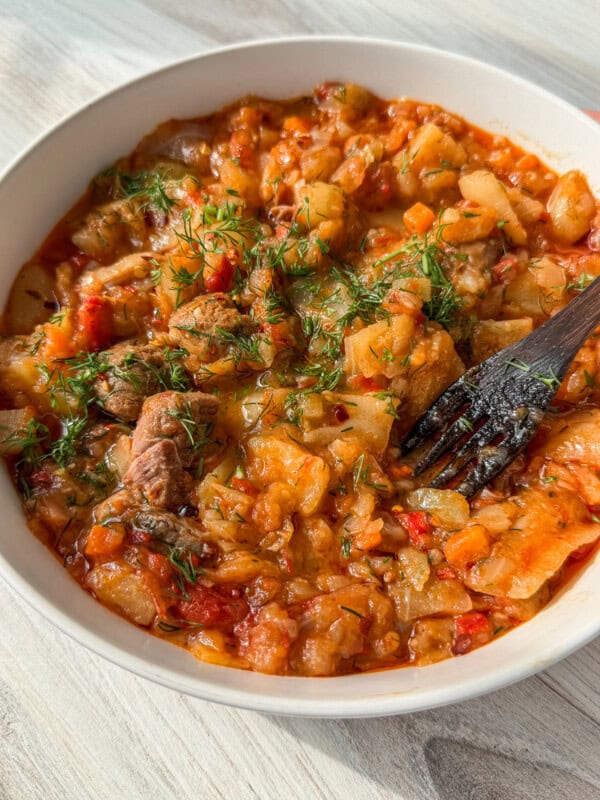 A close-up of a bowl filled with a hearty vegetable and meat stew, topped with fresh chopped herbs. A wooden fork rests inside the bowl, ready for eating. The dish appears thick, with visible chunks of meat, cabbage, carrots, and tomatoes.