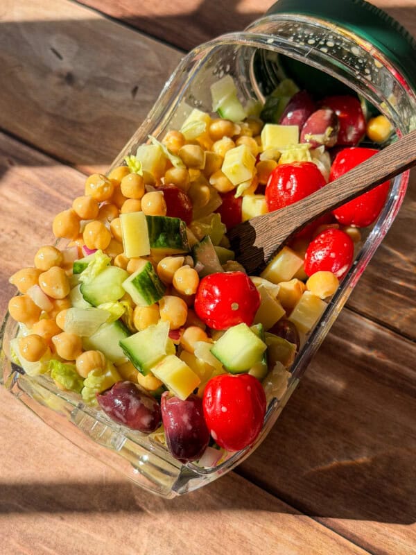 A glass container tipped on its side spills a colorful salad with chickpeas, grape tomatoes, cucumber, cheese cubes, and olives onto a wooden surface. A wooden fork rests inside the container.