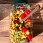 A jar filled with a colorful salad of cherry tomatoes, chickpeas, cucumbers, cheese cubes, and olives sits on a wooden surface, with sunlight casting shadows and a wooden spoon inside the jar.