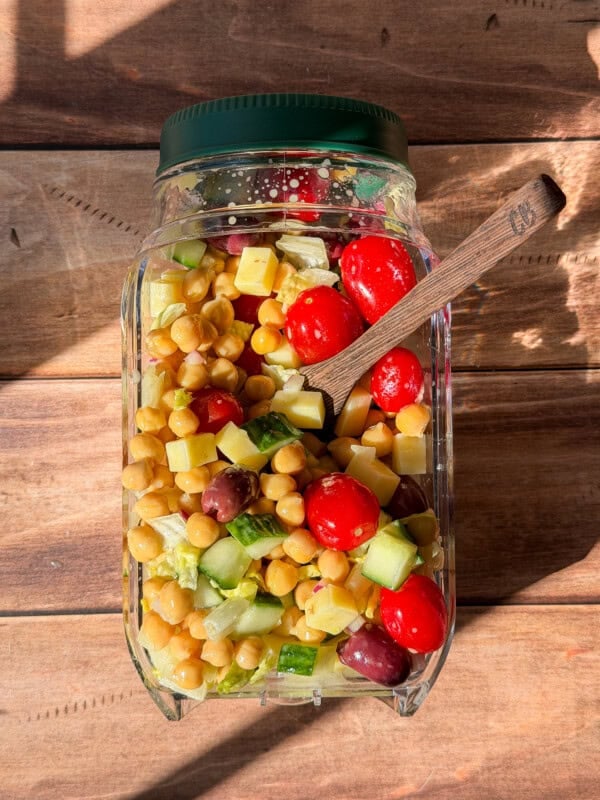 A jar filled with a colorful salad of cherry tomatoes, chickpeas, cucumbers, cheese cubes, and olives sits on a wooden surface, with sunlight casting shadows and a wooden spoon inside the jar.