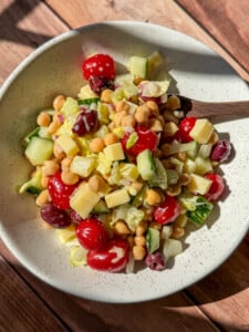 A bowl of salad with cherry tomatoes, chickpeas, kidney beans, chopped cucumber, lettuce, and small cubes of cheese, served in a white speckled bowl on a wooden surface.