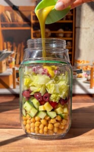 A hand pours yellow salad dressing into a glass jar filled with layered chickpeas, cucumbers, olives, lettuce, and diced red onions. The jar sits on a wooden table in a sunlit kitchen.
