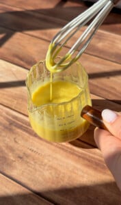 A hand holds a measuring cup of yellow sauce for a Greek Salad Jar while another lifts a whisk, letting the sauce drip back into the cup on a wooden surface in bright sunlight.