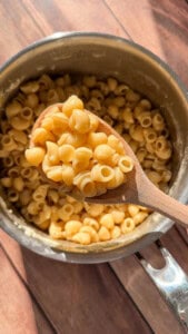 A wooden spoon holds a serving of cooked macaroni shells above a pot filled with more rich, Asian Italian Buttered Noodles, sitting on a wooden surface with sunlight shining on it.
