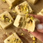 A hand holds a square piece of pistachio fudge with a bite taken out of it, reminiscent of Pistachio Yogurt Bites. More pieces topped with chopped pistachios are scattered on parchment paper in the background.