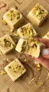 A hand holds a square piece of pistachio fudge with a bite taken out of it, reminiscent of Pistachio Yogurt Bites. More pieces topped with chopped pistachios are scattered on parchment paper in the background.