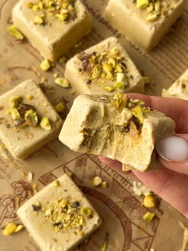 A hand holds a square piece of pistachio fudge with a bite taken out of it, reminiscent of Pistachio Yogurt Bites. More pieces topped with chopped pistachios are scattered on parchment paper in the background.