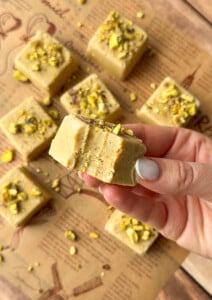 A hand holds a square piece of pistachio fudge with a bite taken out of it; more Pistachio Yogurt Bites topped with chopped pistachios are arranged on brown parchment paper in the background.