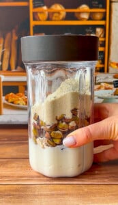 A close-up of a blender cup filled with yogurt, pistachios, and protein powder—perfect for Pistachio Yogurt Bites—held by a hand with light pink nail polish. In the background, baked goods sit on shelves above a wooden table.