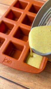 A thick yellow batter for Pistachio Yogurt Bites is being poured from a bowl into the square cavity of an orange silicone mold tray labeled "joie," set on a wooden surface.