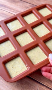 A hand holds a brown silicone mold with nine square cavities filled with a light yellow, speckled mixture, likely Pistachio Yogurt Bites, on a wooden surface.