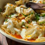 A close-up of Viral Blanket Dumplings topped with creamy yogurt, chopped herbs, and a drizzle of red chili oil in a shallow dish, with a wooden spoon resting on the edge. Sunlight highlights the textures and colors.