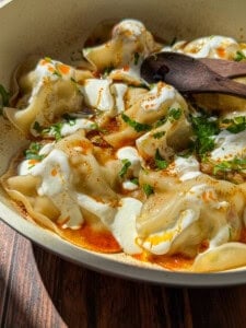 A close-up of Viral Blanket Dumplings topped with creamy yogurt, chopped herbs, and a drizzle of red chili oil in a shallow dish, with a wooden spoon resting on the edge. Sunlight highlights the textures and colors.